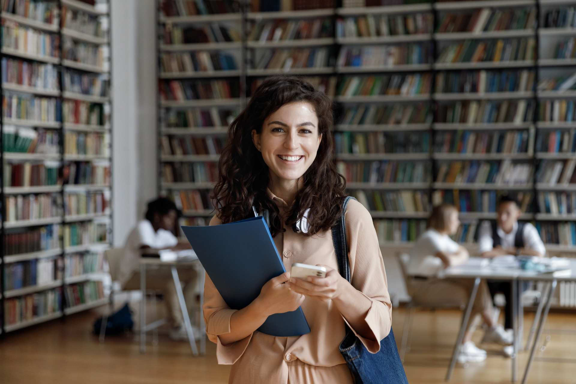 Eine Frau steht in einer Bibliothek. Sie lächelt und hält ein Buch und ein Smartphone in der Hand.
