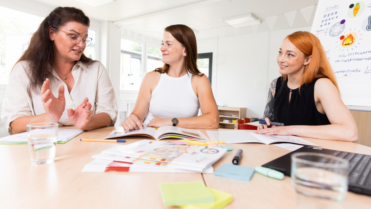 Drei Frauen sitzen an einem Tisch und sprechen über die Unterlagen, die vor ihnen auf dem Tisch ausgebreitet liegen. 