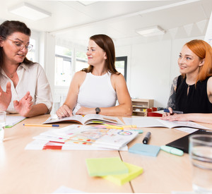Drei Frauen sitzen an einem Tisch und sprechen über die Unterlagen, die vor ihnen auf dem Tisch ausgebreitet liegen.