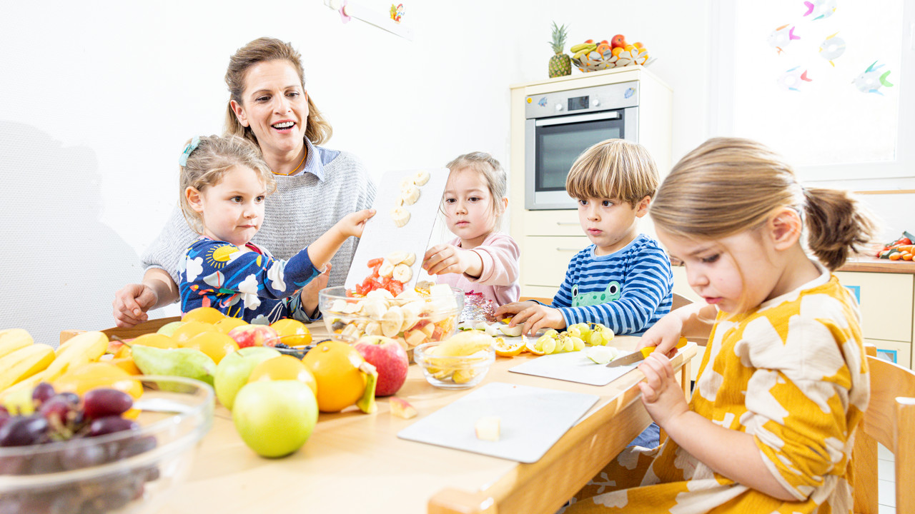 Kinder helfen einer Fachkraft bei der Zubereitung eines Obstsalats.