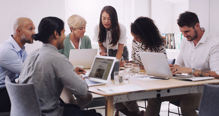 Mitarbeitende sitzen um einen Tisch, zum Teil an Laptops, und arbeiten gemeinsam