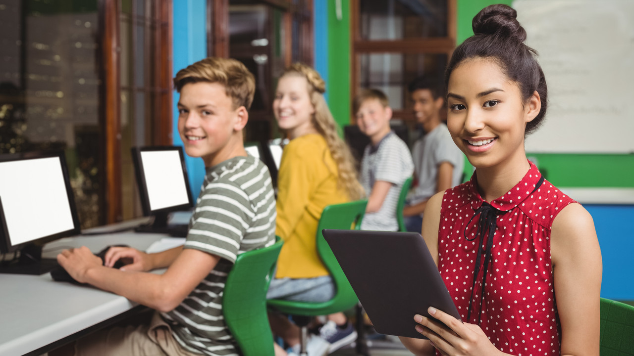 Ältere Schülerinnen und Schüler die jeweils an einem eigenen Computer sitzen. Eine Schülerin steht im Vordergrund, lächelt in die Kamera und hält ein Tablet in der Hand. 