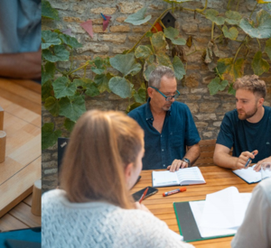 Links: Holzfiguren stehen auf einer Holzplatte, eine wird von einer Hand bewegt; Rechts: vier Erwachsenen sitzen an einem Tisch und diskutieren über etwas.