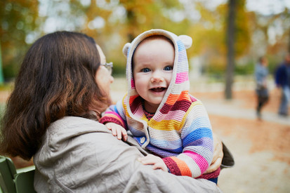 Frau hält ein lächelndes Kleinkind in einer bunten Strickjacke auf dem Arm.
