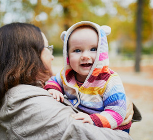 Frau hält ein lächelndes Kleinkind in einer bunten Strickjacke auf dem Arm.
