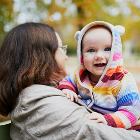 Frau hält ein lächelndes Kleinkind in einer bunten Strickjacke auf dem Arm.